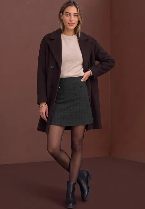 Femme en manteau marron, pull, mini-jupe à carreaux, collants noirs et bottines sur fond marron.