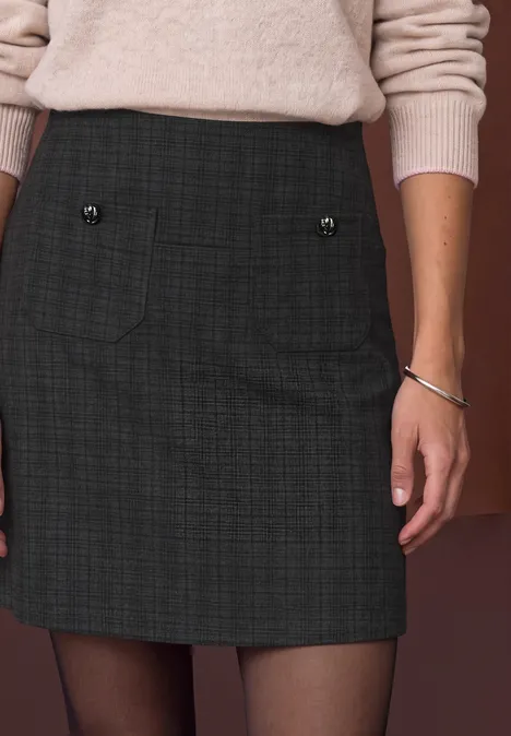Femme portant une minijupe courte à carreaux avec poches boutonnées, pull clair et bracelet argenté.