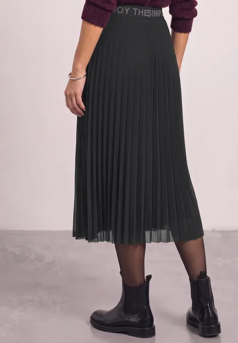 Seitliche Ansicht einer Person in schwarzem, plissiertem Midirock, schwarzen Stiefeletten und Strumpfhose.