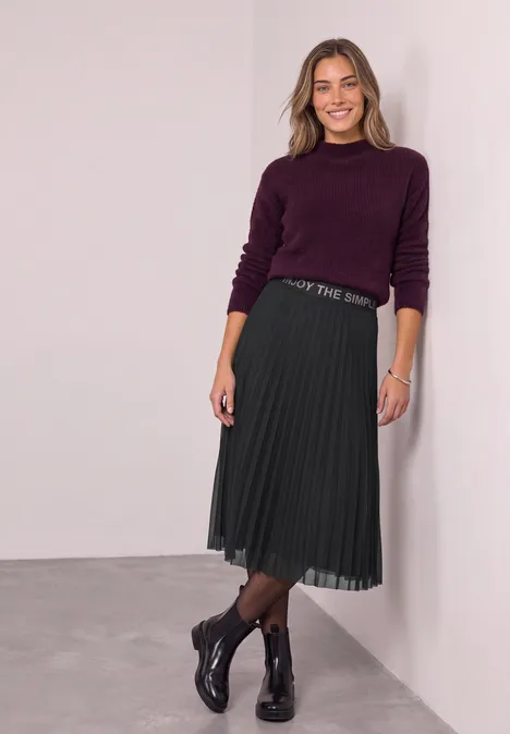 Lächelnde Frau in violettem Pullover, Plissee-Midirock und Stiefeletten lehnt an weißer Wand.