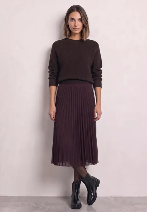 Frau in dunkelbrauem Pullover, plissiertem Midi-Rock und schwarzen Stiefeln vor hellem Hintergrund.