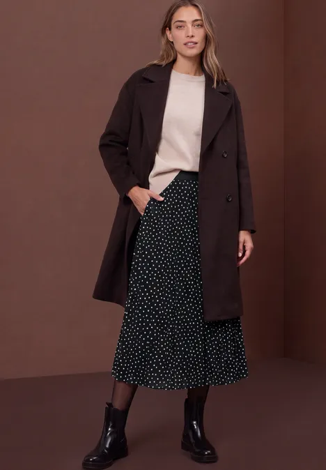 Femme en manteau brun foncé, pull clair, jupe midi noire à pois et bottines noires devant un fond marron.