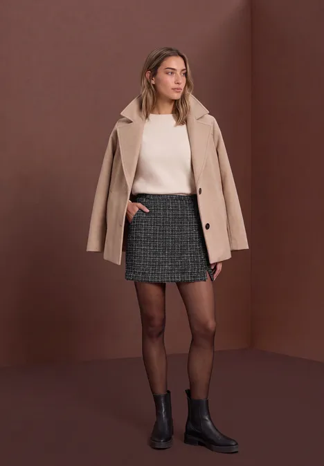 Femme en manteau beige, pull clair, mini-jupe à carreaux, collants noirs et bottines.