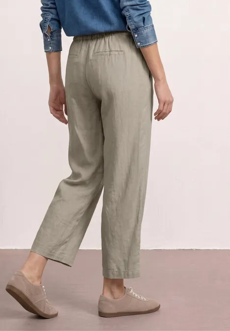 Achteraanzicht van een persoon met kaki linnen broek tot op de enkels, elastische taille en beige veterschoenen.