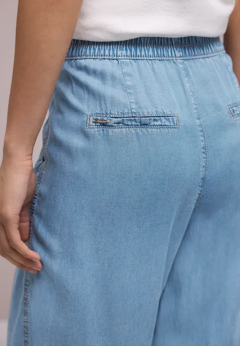 Achteraanzicht van een persoon in lichtblauwe, losse denimbroek met elastische tailleband, hand langs het lichaam.