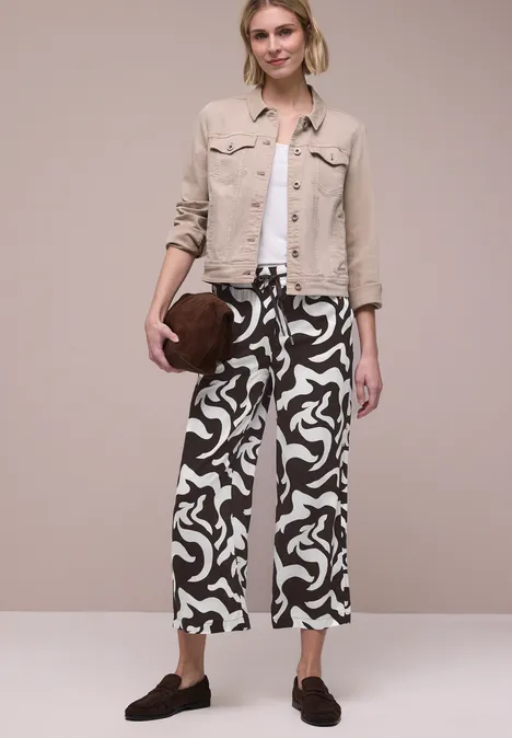 Vrouw met beige spijkerjack, witte top en zwart-witte abstracte broek, bruine ronde clutch en loafers.