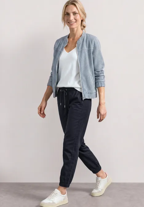 Vrouw in casual outfit: lichte bomberjas, wit t-shirt, donkere joggings en witte sneakers.