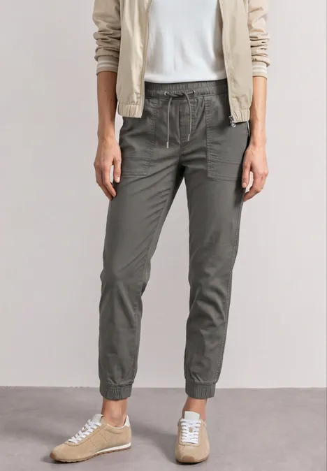 Frau trägt weiße Bluse, helles Jackett, olivgrüne Jogginghose und beigefarbene Sneaker.