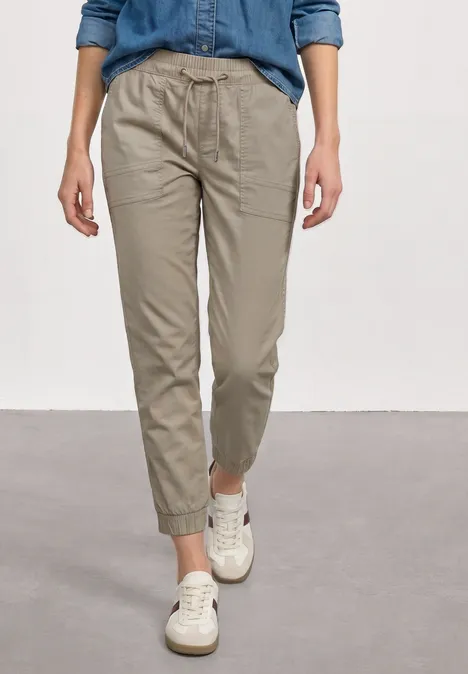 Slim Leg broek in papertouch kwaliteit buff grey Persoon in beige chino met elastische taille, opgerolde pijpen en witte sneakers voor een grijze achtergrond