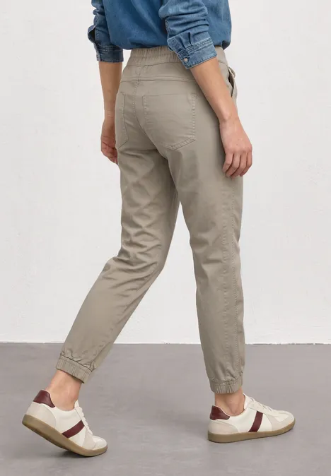 Slim Leg broek in papertouch kwaliteit buff grey Persoon van achteren in beige joggingsbroek en witte sneakers, blauwe spijkershirt tegen witte muur.