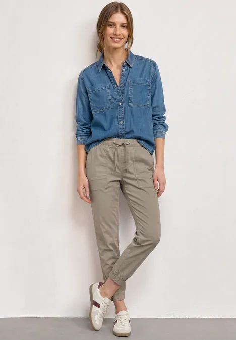 Slim Leg broek in papertouch kwaliteit buff grey Vrouw in jeansoverhemd, beige chino en witte sneakers tegen lichte achtergrond.