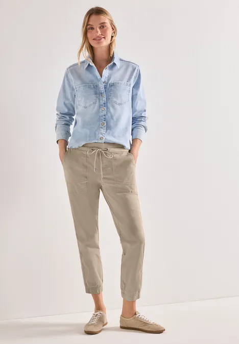 Slim Leg Hose in Papertouch-Qualität cobble stone Frau trägt hellblaues Jeanshemd, beige Jogginghose und Sneakers, steht vor hellem, neutralem Hintergrund.