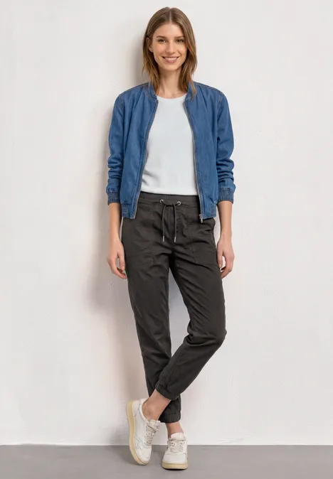 Slim Leg Hose in Papertouch-Qualität deep earth Frau in lässigem Outfit mit blauer Jacke, weißem T-Shirt, schwarzen Hosen und weißen Sneakern vor neutralem Hintergrund.