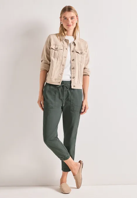 Slim Leg Hose in Papertouch-Qualität cilantro green Frau vor neutralem Hintergrund, trägt beige Jacke, weißes Shirt, grüne Jogginghose und Slip-ons.