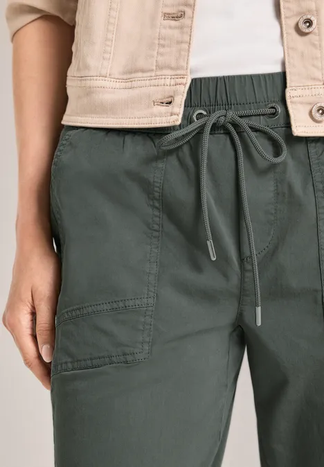 Slim Leg Hose in Papertouch-Qualität cilantro green Nahaufnahme einer dunkelgrünen Hose mit Gummibund und Kordel, Teil einer hellen Jacke sichtbar.