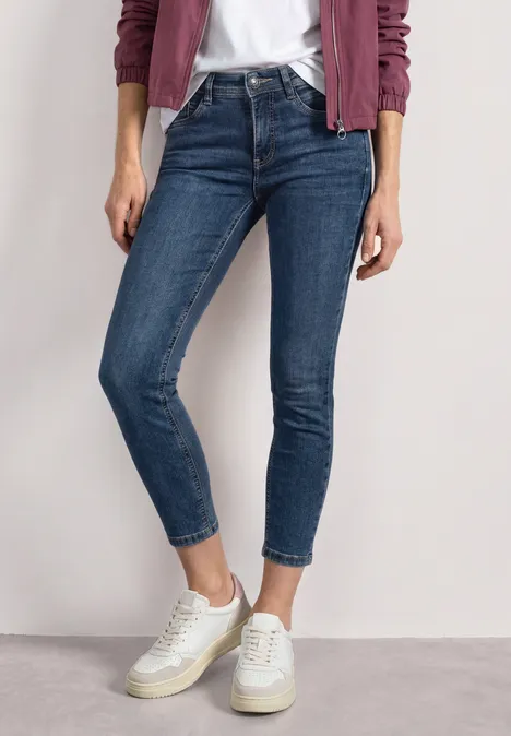 Mid Waist Slim Leg Jeans im Slim Fit brilliant indigo random wash Eine Person trägt schmale blaue Jeans und weiße Sneaker, kombiniert mit weißem Shirt und rosa Cardigan.