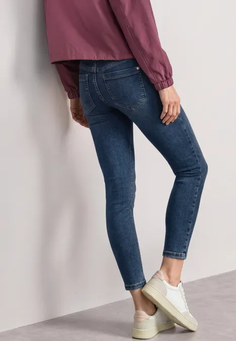Mid Waist Slim Leg Jeans im Slim Fit brilliant indigo random wash Person in engen blauen Jeans, weißem Sneaker und lila Oberteil, seitlich an Wand gelehnt.