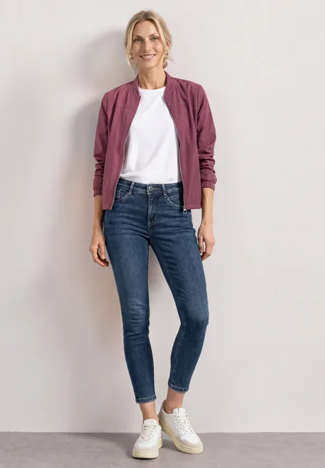 Mid Waist Slim Leg Jeans im Slim Fit brilliant indigo random wash Frau in weißem T-Shirt, bordeauxfarbener Jacke, blauen Skinny Jeans und weißen Sneakern vor hellem Hintergrund.