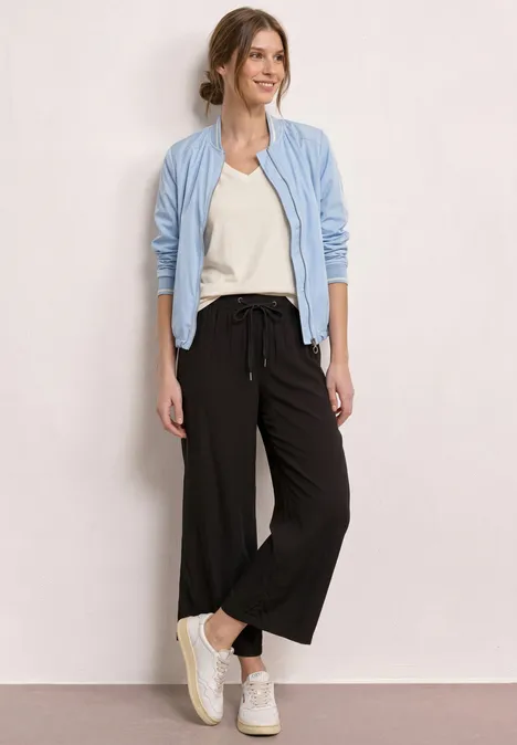 Vrouw met lichtblauw vest, wit shirt, zwarte wijde cropped broek en witte sneakers tegen een neutrale achtergrond.