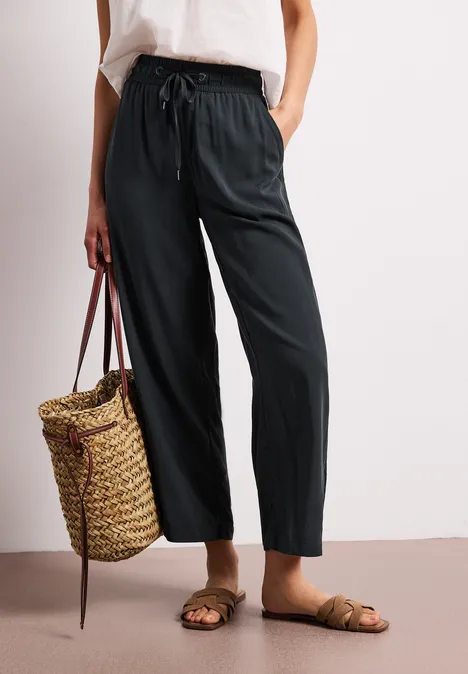 Een persoon draagt een losse zwarte broek, sandalen en houdt een gevlochten strooien tas vast.