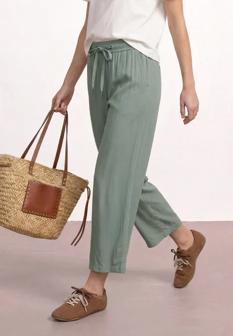 Persoon draagt een losse groene broek, wit t-shirt, bruine schoenen en een strooien tas.