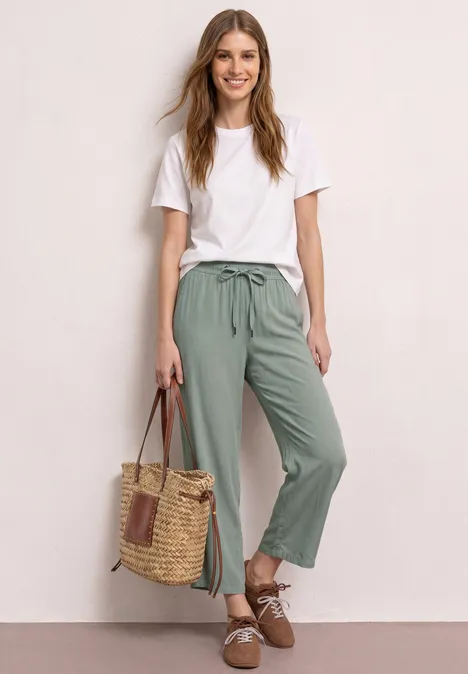 Vrouw leunt tegen muur, draagt wit t-shirt, groene broek, rieten tas en bruine schoenen.