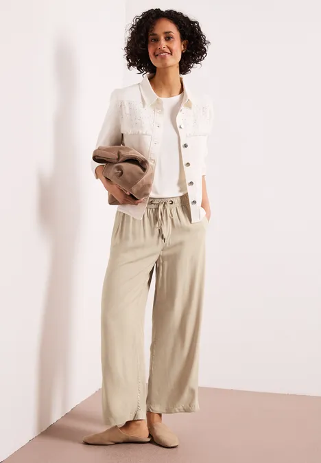 Eine Frau in weiten beigen Leinenhosen, weißer Jacke und beige Loafers, sie hält eine braune Jacke.