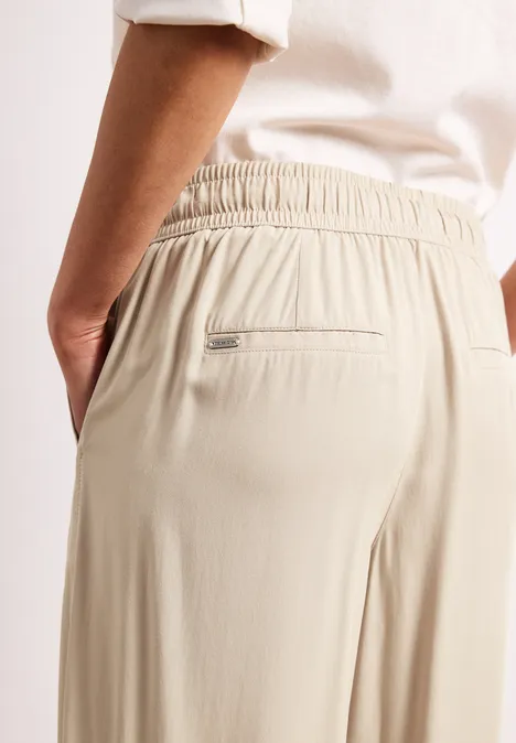 Person trägt beige lockere Hose mit Gummibund und Reißverschlusstasche am Rücken, Hand in der Seitentasche.