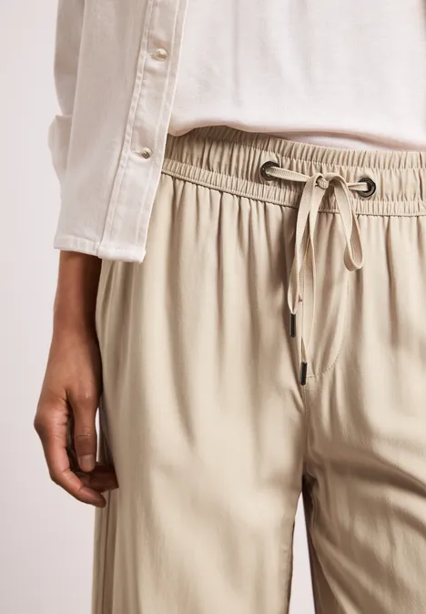 Person in beigefarbener Stoffhose mit Tunnelzug und weißem Oberteil, Hand an der Seite, vor hellem Hintergrund.