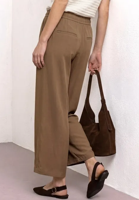 Frau von hinten in weiten, braunen Hosen, weißem Top, schwarzen Sandalen und brauner Schultertasche.