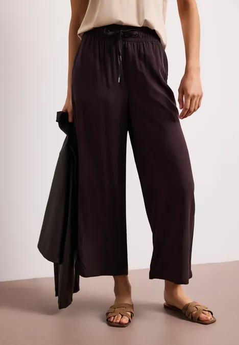 Locker geschnittene, schwarze Stoffhose mit Tunnelzug, kombiniert mit braunen Sandalen und passender Jacke in der Hand.