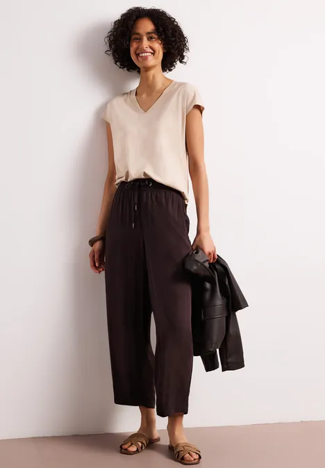 Frau mit lockigem Haar in beigem Top und weiter schwarzer Hose, hält schwarze Lederjacke.