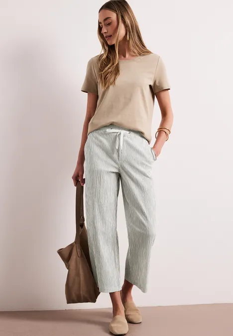 Frau in beigem T-Shirt, hellgrünen gestreiften Culottes, beigen Loafers und Tasche vor neutralem Hintergrund.