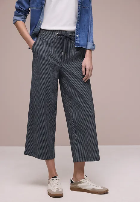 Dame in weißem Shirt, Jeansjacke und weiten, gestreiften 3/4-Hosen mit Taschen dazu weiße Slipper.