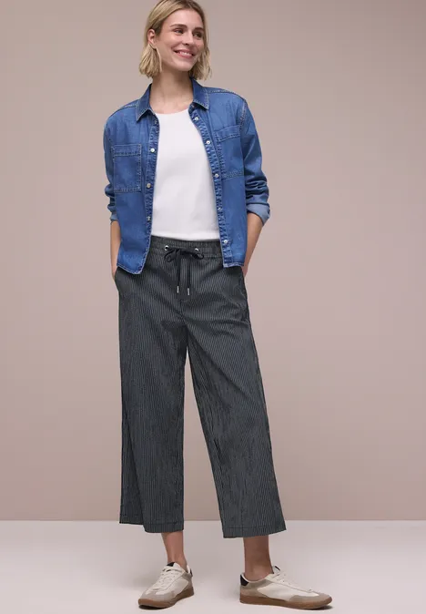 Frau mit Jeanshemd, weißem T-Shirt und weiten grauen Hosen vor beigem Hintergrund.