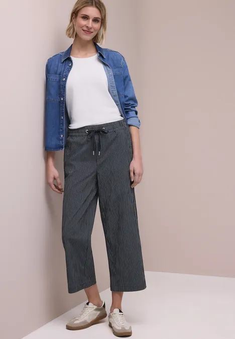 Eine Person in Grauhose, weißem T-Shirt, Jeanshemd und weißen Sneakern steht vor einer beigen Wand.