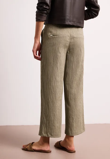 Rückansicht einer Person in locker sitzenden, plisseeartigen olivgrünen Culotte-Hosen und flachen Sandalen.