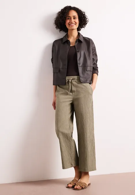 Frau mit lockigem Haar trägt schwarzen Pullover, dunkelgraue Jacke, olivgrüne weite Hose und Sandalen vor hellem Hintergrund.