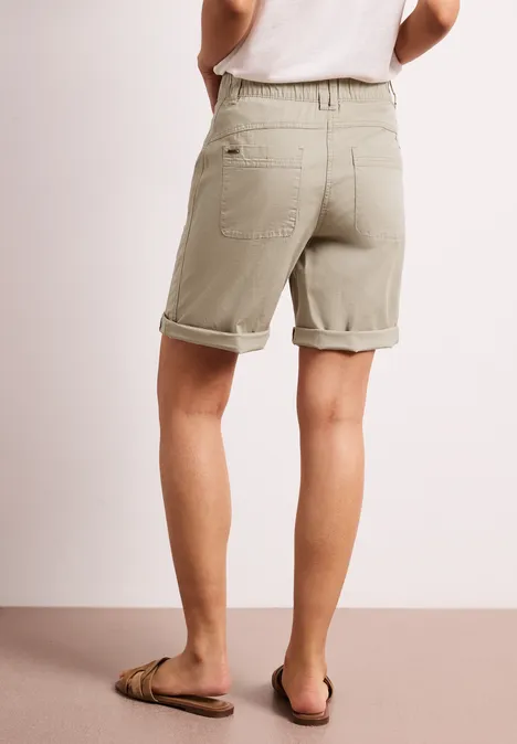 Rückansicht einer Person in khakifarbenen, aufgekrempelten Shorts und Flip-Flops vor hellem Hintergrund.