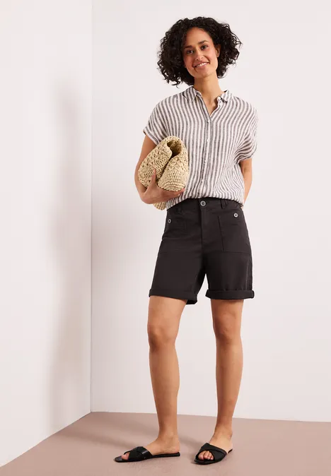 Persoon in een hoek van de kamer draagt gestreept overhemd, zwarte shorts en sandalen, met strooien tas.