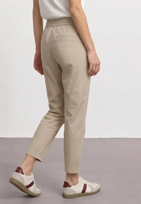 Zijaanzicht van een persoon in beige, casual broek en lichte sneakers tegen een neutrale achtergrond.