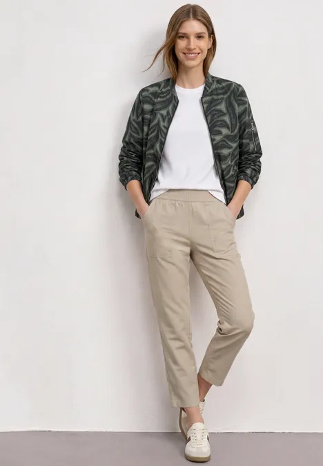 Vrouw in wit T-shirt, groene jas met bladerenprint, beige broek en witte sneakers, staand voor een witte muur.