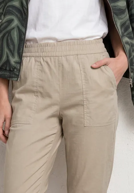 Beige broek met elastische taille en opgestikte zakken, hand ontspannen in de zak gestoken.