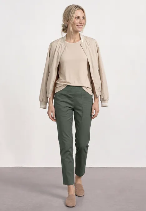 Een vrouw draagt een beige jas, een beige shirt, donkergroene broek en beige schoenen voor een neutrale achtergrond.