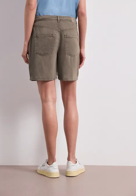Person von hinten in khakifarbenen Shorts und weißen Turnschuhen, stehend vor neutralem Hintergrund.