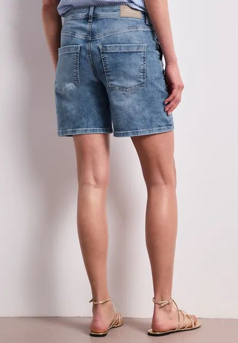 Rückansicht einer Person in Jeansshorts und flachen Sandalen vor weißem Hintergrund.