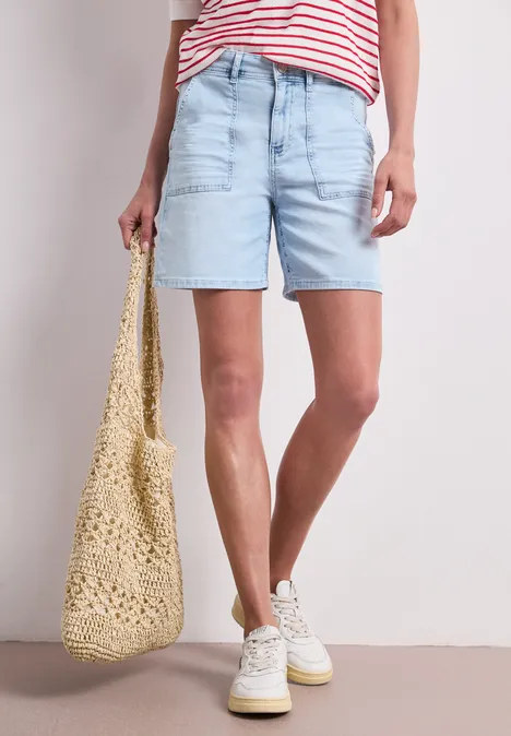 Frau in hellblauen Jeansshorts, gestreiftem Shirt, weißen Sneakern und Häkel-Tasche.