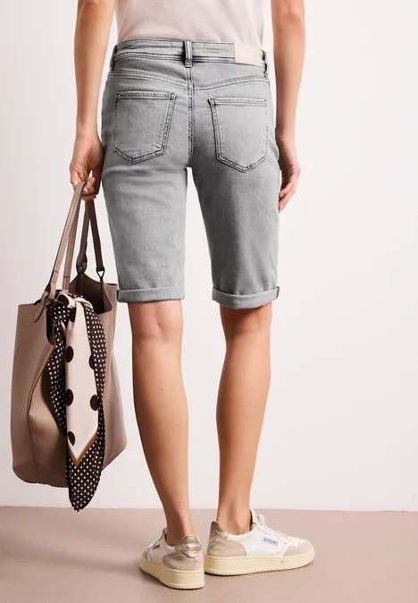 Person von hinten in hellgrauen Denim-Bermuda-Shorts, trägt beige Sneaker und eine tote Tasche mit gepunktetem Tuch.
