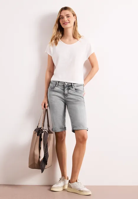 Frau in weißem T-Shirt, grauen Denim-Bermudas und Sneakern mit großer Tasche.