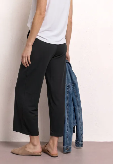 Vrouw met wit topje en zwarte wijde culotte, houdt een spijkerjasje vast en draagt slippers.